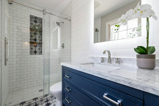A modern bathroom with a blue vanity, white marble countertop, and a glass shower enclosure. The wall features a white tile design with a decorative pattern near the shower.
